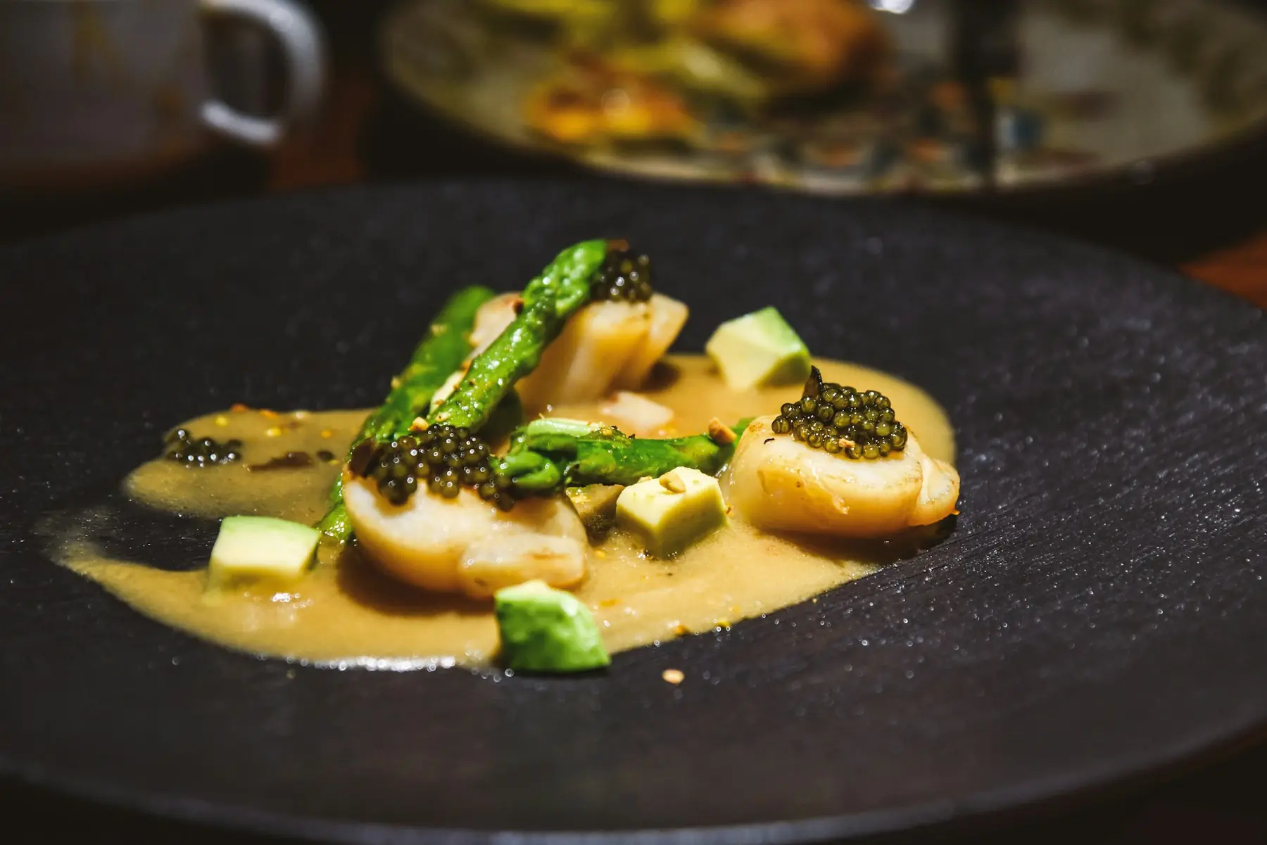 Gebratene Jakobsmuscheln mit grünem Spargel, Kaviar und heller Sauce auf dunklem Teller